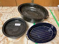 Set of three enamelware roasting pans displayed on countertop: two large black speckled oval pans (one with lid) and one small blue speckled round pan with metal rack.