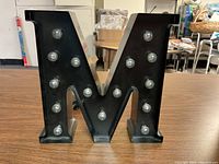 Front view of a black metal letter M with clear bulbs arranged for illumination.