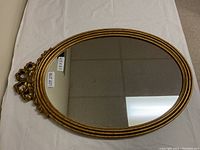Oval gold framed wall mirror with decorative crest laying flat on white cloth showing tags with size 19 x 28 and lot number
