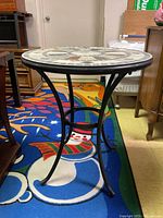 Photo of the full oval mosaic tile top occasional table with black curved metal legs positioned on a colorful rug with a snowman design.
