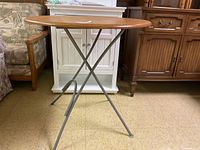 Side view of round folding table showing wooden top and metal X-frame legs on a tiled floor in a room with other furniture.