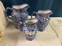 Photo showing three floral ceramic jugs in varying sizes with blue, pink, and yellow flower decoration on a white background.