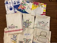 Overview of ten vintage tea towels folded and arranged on a wooden surface. Display includes embroidered floral, kitchen motif designs, and printed floral patterns.