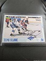 Front of the Teemu Selanne autographed hockey card showing Selanne in white and blue uniform skating during a game moment with a black ink autograph across the card.