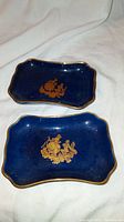 Pair of small Limoges porcelain dishes, cobalt blue with gold designs inside and gold trim on edges, shown from above on white cloth.