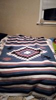 Photo showing full view of handmade Mexican blanket with central Aztec motif on a bed, displaying colors and pattern details including stripes and geometric design