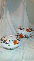 Two Royal Worcester Imari Grainger casseroles with lids, showing floral and ribbon patterned décor in orange, blue, and green, displayed on white fabric.