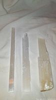 Three selenite gemstone wands arranged on a light cloth background, showing their translucent white to beige coloration and natural striations. The wands vary in height and have some rough edges.