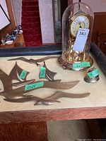 Anniversary clock under glass dome with numbered tag and brass wall art pieces with size labels on tabletop.