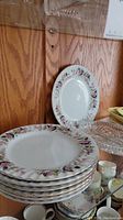Photo showing stacked dinner plates with floral rose pattern and gold trim on edges, displayed on a glass shelf with wooden back.