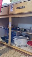 Photo shows under-shelf kitchen cabinetry with various cookware including multiple saucepans, a white Crockpot with glass lid, and a few other kitchen tools visible on shelves.