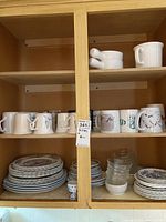 Photo showing two shelves of dishes, including stacked plates, various mugs, and some small bowls and glasses.