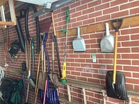 Various garden tools hanging on brick wall including dustpans, shovels, rakes, wrenches, watering can, and extension ladder.