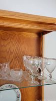 Shelf with clear glassware including several stemmed glasses, creamer, sugar bowl, ashtray, and candle holders visible on glass shelf in wooden cabinet