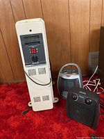 Photo of three heaters: tall white Holmes Air heater with black control panel, small silver Alpina heater, and black Classic heater with two control knobs. All placed on carpet against wood panel wall.