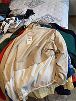 Stack of men's jackets in beige, cream, green and dark colors, laid on a bed, showing different styles and sizes.