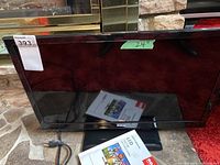 Front view of 24-inch RCA LED TV with power cord and user manual placed on the floor against stone background.