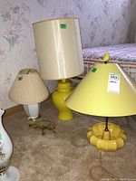 Three lamps assembled on carpeted floor: small white ceramic lamp with beige pleated shade, medium yellow ceramic lamp with large cream drum shade, and yellow lamp with conical shade and bird finial.