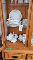 View of the china set inside a glass cabinet showing stacked plates of various sizes, a vegetable bowl on top, and the teapot, sugar bowl, creamer, gravy boat, and cups with saucers arranged below.