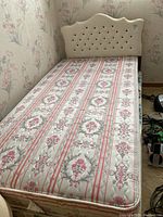 Single bed with tufted cream headboard, floral stripe mattress on top, and floral box spring beneath. Electrical items and other small objects visible alongside.