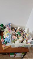 Wide angle view showing assortment of small spring and Easter-themed figurines arranged in a plastic bin and wooden base display with flower shapes.