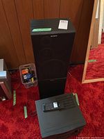 Two black Sony speakers stacked vertically with a plastic box of assorted batteries next to them, all on red carpet
