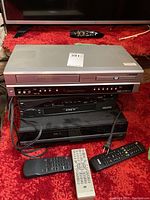 Three stacked electronic media players with three remote controls on red carpet under TV.