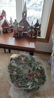 Photo showing four Christmas village buildings arranged on a wooden table near window with snowy background and a decorative wreath wrapped in plastic on the floor nearby.