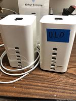 Two white Apple AirPort Extreme WiFi routers shown side by side on a table. Both have multiple Ethernet ports on the back. One unit has a blue tape labeled 'OLD'.