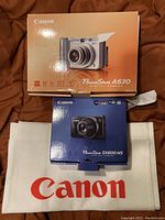 Two Canon PowerShot camera boxes (A630 and SX600 HS) lying on a surface with a Canon branded white pouch beneath them.