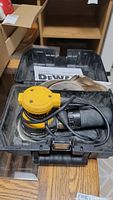 Yellow and black Dewalt 1/4 sheet sander in black hard case with coiled power cord and visible sanding pad inside. Instruction manual also seen inside case.