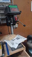 Front angle showing Skil drill press on wooden bench with manuals on base.
