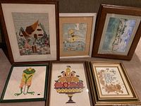 All six framed artworks displayed together on carpeted floor showing varied sizes and styles including embroidery, collage, golf art, watercolor, fruit print and owl embroidery.