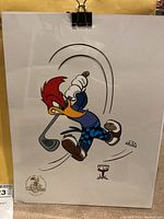 Full view of the lithograph print showing Woody Woodpecker swinging a golf club, displaying the entire artwork and the seal in the bottom left corner.