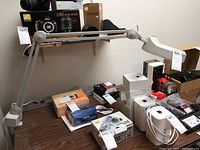 Clamp-on magnifying desk lamp positioned on a table with various electronics boxes in the background.