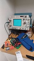 Image of vintage 465 oscilloscope set up on table with screen and visible knobs, digital multimeter and blue carrying case placed on table surface below oscilloscope.