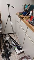 Silk 500G tripod with adjustable head, legs, and green locking clips standing upright by a wall with other tripods on the floor nearby