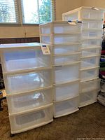 Three clear plastic rolling storage cabinets shown standing side-by-side in room with carpet and wood paneling. Each cabinet includes multiple drawers and wheels.