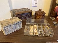 Photo showing two carved wood boxes, one round and one rectangular, one rectangular marble box, and a plastic organizer with shells and earrings.