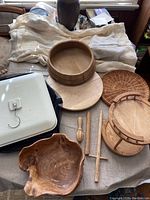 Overall lot on table showing large burl wood bowl, round wooden bowl, two-tier serving dish, rattan tray, basket, wooden utensils, and white square tray.