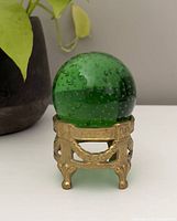 Green glass sphere resting in brass holder on white surface next to plant pot.