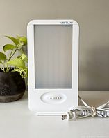 Front view of Verilux HappyLight showing frosted light panel, front switch, power cord coiled beside it, next to a plant.