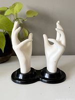 Front view of two white ceramic display hands on a black base, positioned with plants in background