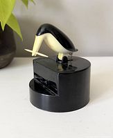 Front angled view showing black bird figure holding a toothpick on circular base.