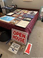 Large assortment of framed items, signs, and posters laid out on a table and floor, showing various sizes and styles