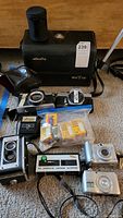 Photo showing all cameras and accessories together including Kodak Duaflex II, Minolta cameras, Nikon and Canon digital cameras, Vivitar flash, and Kodak film rolls in packaging.