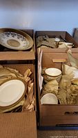 Boxes containing porcelain china including plates, bowls, teacups, saucers, and a gravy boat, wrapped in protective paper.