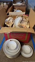 Boxes filled with various china dishes wrapped in newspaper, stacked plates on floor with visible golden pine designs.