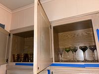 Top left and right open wooden cabinets show amber glasses on the left and colored aperitif glasses on the right shelf.