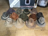 Overview photo showing 13 men's felt fedora hats in various earth tone colors, arranged on a wooden floor.